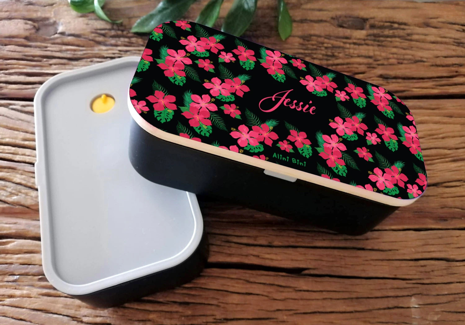 A Munch-Mate lunch box, lying on a wooden table, showing a black and pink floral print, personalised with the name Jessie
