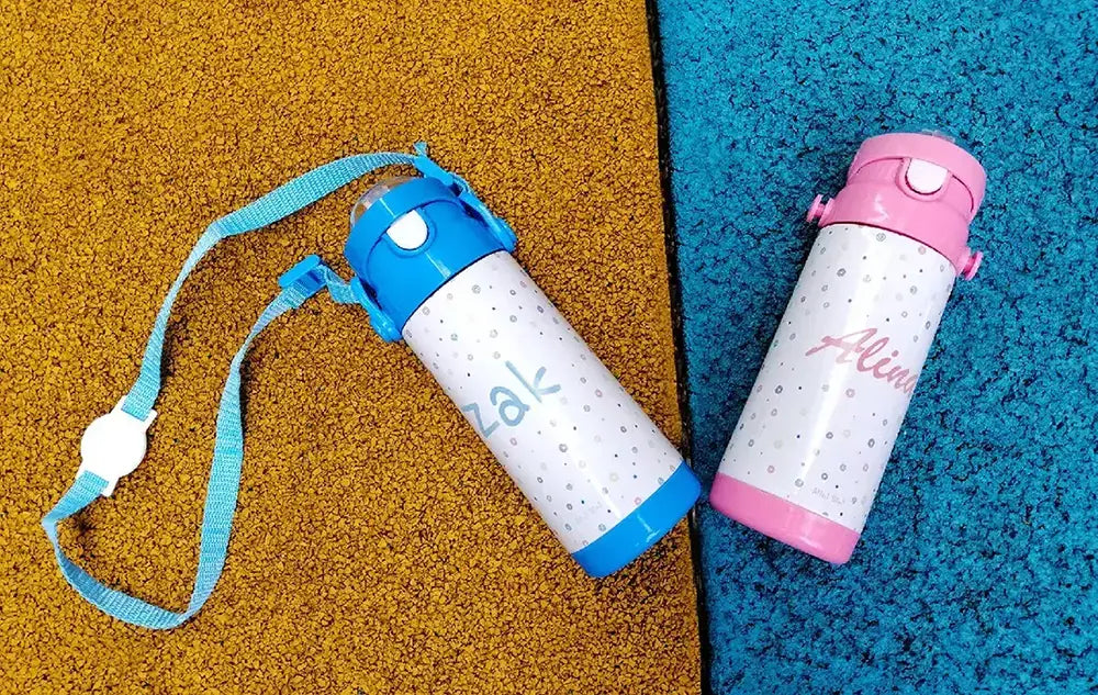 Two personalised Sip-n-Go bottles lying on a playground floor, one blue and one pink.